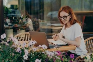 Redhead skillful woman student writes in notebook sits in front of opened laptop computer photo