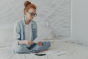 Young woman sits crossed legs on comfortable bed checks financial documents calculates family budget photo