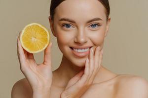 Close up shot of beautiful tender woman smiles gently has healthy well groomed skin holds slice of lemon uses citrus fruit for beauty and health stands shirtless isolated over brown background