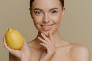 Cropped shot of adorable smiling female model has tender skin touches face gently holds yellow juicy lemon uses natural cosmetic stands topless indoor against brown background. Health food concept