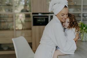 Happy affectionate young mother in bath towel and dressing gown embraces with love who sits at table feel refreshed after taking shower spend free time at home. Family beauty wellness concept