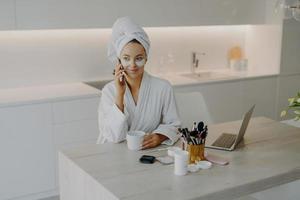 Lovely female model wears bathrobe and soft towel wrapped on head undergoes skin care procedures applies beauty patches under eyes has coffee break while talking with friend via cellular being at home