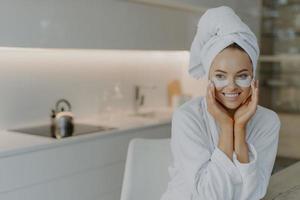 Photo of lovely woman with healthy skin applies recovery pads under eyes enjoys skin care and effective beauty treatment dressed in soft bathrobe wears wrapped towel on head poses over home interior