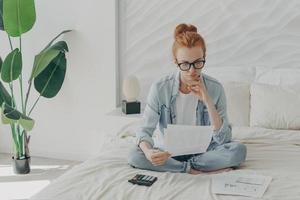 mujer europea de jengibre enfocada sentada en la cama y calculando facturas domésticas en casa, sosteniendo papeles foto