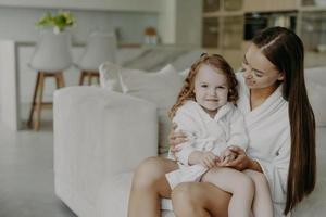 Beautiful brunette affectionate mother holds small curly haired cute dauhter in dressing gown pose in cozy room against modern apartment interior. Mom and little girl at home after taking shower