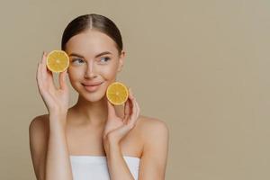 Horizontal shot of thoughtful young European woman uses homemade fruit for facial mask holds lemon slices wrapped in white soft bath towel looks aside isolated over brown background copy space photo