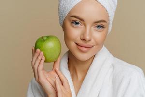Close up shot of blue eyed woman with minimal makeup well cared complexion fresh clean skin dressed in white coat holds green apple isolated over brown background. Natural cosmetics concept. photo
