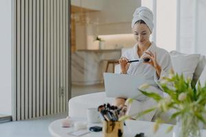 Lovely young Caucasian woman wears bathrobe and towel on head holds cosmetic brush in front of laptop computer gives tutorail lesson how to do makeup sits at comfortable sofa in modern apartment photo