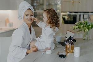 Pretty small girl with curly hair looks how mother does cosmetic procedures applies collagen patches under eyes for skin treatment wear bathrobes pose near desk with beauty products. Everyday care photo