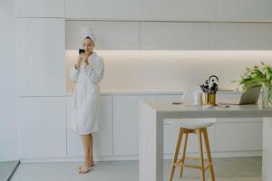 Full length shot of young beautiful woman dressed in white bathrobe applies face cream looks in mirror takes care of her skin poses near kitchen furniture stands bare feet. People beauty facial care