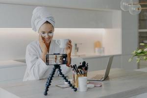 Woman beauty blogger shoots cosmetic vlog looks at camera of smartphone talks with followers applies collagen patches under eyes drinks tea wears white soft bathrobe records video broadcast. photo