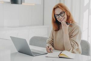 Positive ginger woman in casual clothes records audio message or smartphone while working on laptop photo