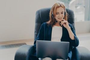 Young attractive european woman wearing headset sitting with laptop on armchair at home office photo