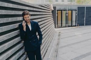 CEO male in having pleasing phone talk on mobile while standing outside of office center entrance