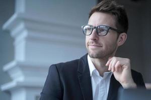 Confident attractive young businessman sitting at home office, looking aside with pensive expression photo