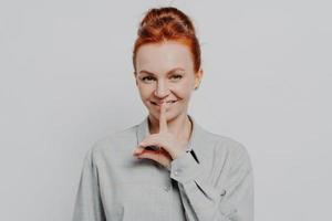 Cheerful young ginger 30s woman showing hush gesture while posing isolated on grey studio background photo