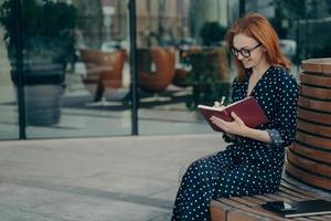 bella y elegante mujer de negocios de jengibre tomando notas mientras se sienta en un banco de madera en el centro de la ciudad foto