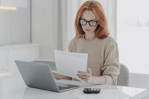 joven mujer pelirroja enfocada sentada en la mesa con una computadora portátil y sosteniendo facturas de impuestos en papel foto