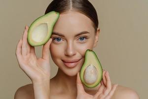 primer plano de una tierna mujer de ojos azules mira directamente a la cámara sostiene mitades de aguacate cerca de la cara elige una nutrición saludable tiene una piel suave perfecta aislada sobre fondo beige. concepto de belleza foto