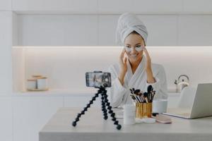 Happy young Caucasian woman records beauty blog on mobile phone standing on tripod goves advice how to use cosmetic patches shoots video wears bathrobe poses at home uses high speed internet photo
