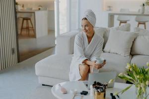 Relaxed thoughtful woman dressed in bathrobe and wrapped towel on head sits on sofa with cup of beverage near table with cosmetic products looks pensive aside poses against cozy home interior