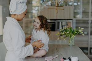 Mother and daughter in white bath robes have pleasant talk with each other going to undergo beauty procedures after taking shower. Small adorable girl with curly hair looks at mommy at home. photo