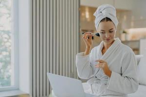 Photo of young European woman applies face powder with cosmetic brush concentrated in laptop computer makes video call has own beauty blog poses in comfortable bathrobe against home interior