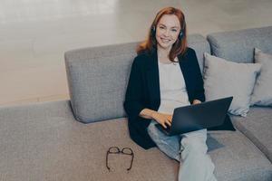 Beautiful smiling woman with red hair sitting with laptop and wireless headset on sofa in office photo