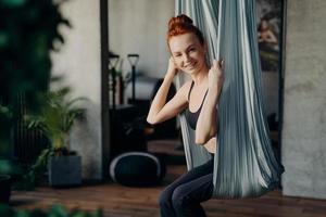 Happy red haired woman sitting in aerial yoga hammock photo