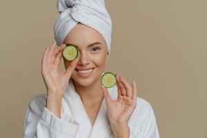 Skin care natural beauty and cosmetology concept. Positive refreshed beautiful woman holds slices of fresh cucumber makes mask for face skin dressed in domestic robe wrapped bath towel on head photo