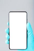 teléfono inteligente con una pantalla blanca limpia en la mano en un primer plano de guante médico azul. foto