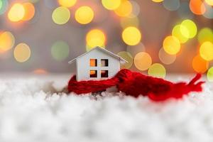 Abstract Advent Christmas Background. Toy model house with knitted red scarf on snow with defocused garland lights background. Christmas with family at home concept. Christmas winter composition. photo