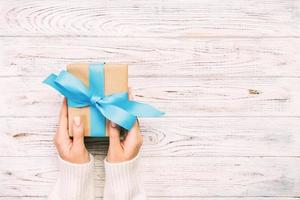 Woman hands give wrapped valentine or other holiday handmade present in paper with blue ribbon. Present box, decoration of gift on white wooden rustic vintage table, top view with copy space photo