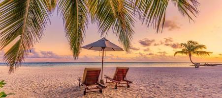 Beautiful tropical sunset beach. Romantic couple goals, two sun beds, umbrella under palm tree leaves. Panoramic coast landscape, love and relaxation banner. Colorful dream travel sea sand sky island photo