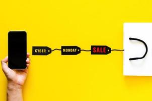 Hand holding smartphone for shopping online with price tags and shopping bag on yellow background. Cyber Monday concept. photo