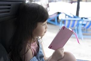 cute little girl sitting in the back of a car reading a pink book near the beach. weekend vacation photo