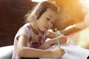 chica asiática sentada en un escritorio en casa escribiendo un libro. educación en línea. nueva normalidad educativa. conceptos de lectura y escritura de los niños. educacion desde casa foto