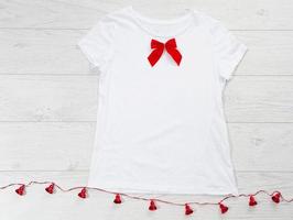 Close up white blank template t shirt with copy space and Christmas Holiday concept. Top view mockup t-shirt and red bow on white wooden background. Happy New Year decorations accessories. Xmas outfit photo