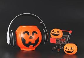 plastic Halloween pumpkin  bucket cover with headphones  isolated  on black  background  with shopping cart or trolley and smaller pumpkins. Halloween holiday shopping  concept. photo