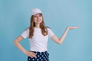 mujer caucásica con camiseta blanca y gorra de fondo azul, apuntando hacia un lado foto