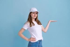 Caucasian woman in white t-shirt and cap on blue background, pointing to side photo