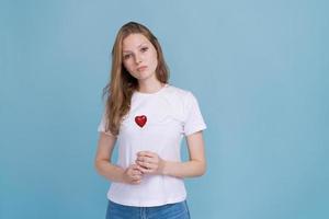 Young woman holds red heart on stick in white t-shirt on blue background. photo