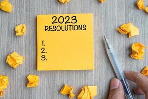 2023 RESOLUTIONS word on yellow note with Businessman holding pen and crumbled paper on wooden table background. New Year start, Strategy and Goal concept photo