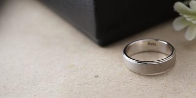 White gold wedding ring with a bird laser on the ring surface. With a black ring box background and artificial flowers. On the left there is an empty space to fill in text photo