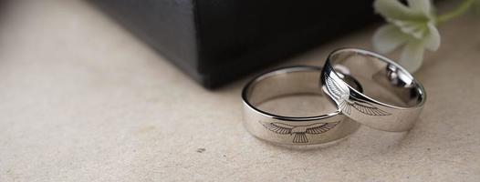 White gold wedding ring with a bird laser on the ring surface. With a black ring box background and artificial flowers. On the left there is an empty space to fill in text photo