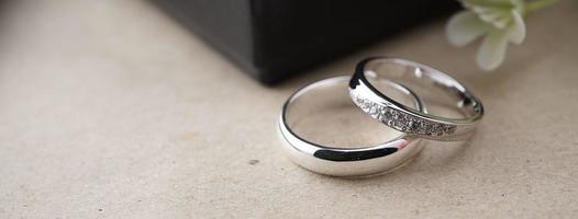 anillo de bodas de oro blanco con un láser de pájaro en la superficie del anillo. con fondo de caja de anillos negra y flores artificiales. a la izquierda hay un espacio vacío para completar el texto foto