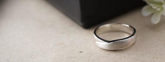 anillo de bodas de oro blanco con un láser de pájaro en la superficie del anillo. con fondo de caja de anillos negra y flores artificiales. a la izquierda hay un espacio vacío para completar el texto foto