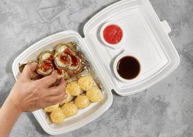 Delicious sushi rolls in disposable plastic boxes. The hand takes a roll with chopsticks. Food delivery service concept, flat stacking photo
