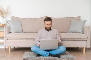 Young attractive man sitting on sofa at home, working on laptop online, using photo