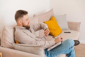 Successful bearded young man writes notes in notebook while sitting at home photo